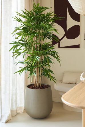 La Plante Artificielle Bambou 150 cm, aux feuilles vertes luxuriantes, est placée à l'intérieur près d'une fenêtre avec des voilages, à côté d'un canapé beige avec un coussin et une décoration murale abstraite dans une pièce moderne décorée dans des tons neutres.
