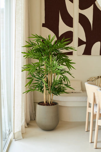 Un bambou artificiel de 120 cm se dresse dans un salon moderne et lumineux près d'une fenêtre avec des voilages, à côté d'une chaise blanche et d'une décoration murale abstraite.