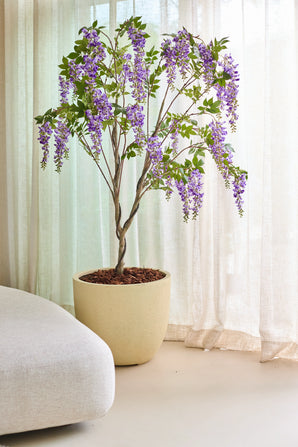 La plante Glycine artificielle 210 cm, avec ses bouquets de fleurs violettes en surplomb, est placée à l'intérieur à côté de rideaux beiges translucides et d'un canapé de couleur claire.