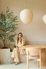 Une femme en costume beige est assise souriante à une table en bois dans une pièce moderne et minimaliste avec des murs clairs, des suspensions en papier suspendues au plafond et une plante d'eucalyptus artificielle de 180 cm qui ajoute un peu de verdure à la pièce.