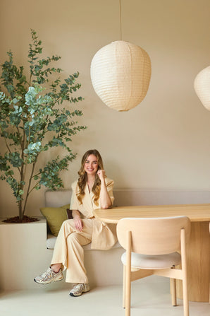 Une femme en costume beige est assise souriante à une table en bois dans une pièce moderne et minimaliste avec des murs clairs, des suspensions en papier suspendues au plafond et une plante d'eucalyptus artificielle de 180 cm qui ajoute un peu de verdure à la pièce.