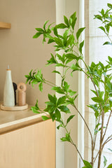 Une plante artificielle Pieris Japonica de 150 cm se dresse près d'une fenêtre à côté d'une armoire en bois clair contenant des bouteilles en céramique et un moulin à poivre, ajoutant une touche minimaliste à la pièce lumineuse et aérée.