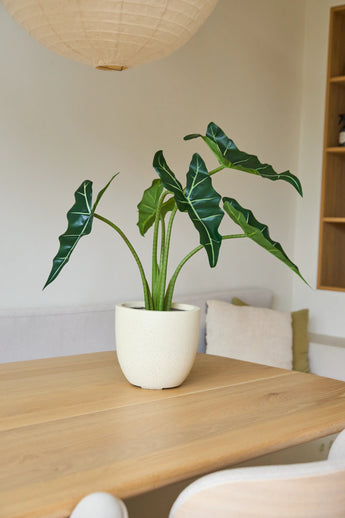 La plante artificielle Alocasia de 80 cm, aux grandes feuilles vertes, se dresse sur une table en bois clair dans une pièce minimaliste à l'éclairage tamisé et à la décoration neutre.