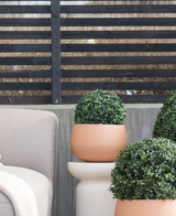 Trois topiaires rondes UV de style romain en buis artificiel de 30 cm se trouvent sur un patio près d'une chaise d'extérieur beige, contre une clôture moderne à lattes noires et des arbres nus.