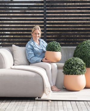 Une femme assise pieds nus sur un banc extérieur de couleur claire, sourit et tient une Plante Artificielle Buis Style Romain UV D30cm. Deux plantes artificielles similaires se trouvent à proximité, avec une clôture en lattes de bois et un décor de patio en arrière-plan.