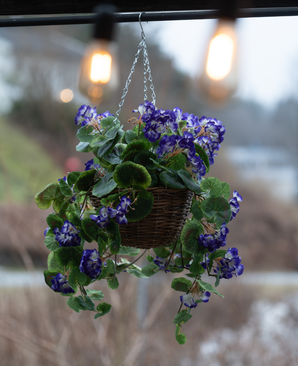 De Kunstbegonia paars in mand UV 70cm is een hanging basket met levendige paarse kunstbegonia's met groene bladeren, perfect voor buiten. Het arrangement voegt kleur en charme toe onder warme gloeilampen.
