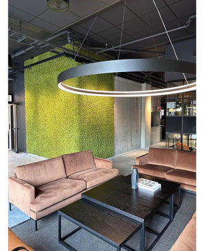 Un coin salon moderne avec des canapés beiges, des tables basses noires, un grand plafonnier rond et un mur vert en arrière-plan, créé avec Kunsthaag Nordic Moss 50x50 cm UV, qui dégage un intérieur luxueux et un design industriel contemporain.