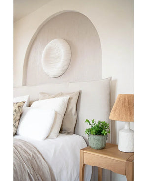 Une chambre confortable avec une tête de lit beige, une literie et des oreillers neutres, une table de chevet en bois surmontée d'une plante artificielle d'eucalyptus de 27 cm, une lampe avec un abat-jour texturé et une décoration murale blanche ronde au-dessus du lit dans une alcôve voûtée.