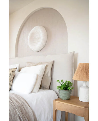 Une chambre confortable avec une tête de lit beige, une literie et des oreillers neutres, une table de chevet en bois surmontée d'une plante artificielle d'eucalyptus de 27 cm, une lampe avec un abat-jour texturé et une décoration murale blanche ronde au-dessus du lit dans une alcôve voûtée.
