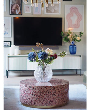 Un salon moderne comprend une table basse en terrazzo avec un vase de fleurs en verre, une télévision au mur, des œuvres d'art et des photos encadrées, un buffet blanc et l'hortensia artificiel Deluxe de 57 cm lilas comme un élégant arrangement de fleurs bleues en arrière-plan.