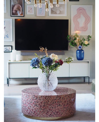 Un salon moderne comprend une table basse en terrazzo avec un vase de fleurs en verre, une télévision au mur, des œuvres d'art et des photos encadrées, un buffet blanc et l'hortensia artificiel Deluxe de 57 cm lilas comme un élégant arrangement de fleurs bleues en arrière-plan.