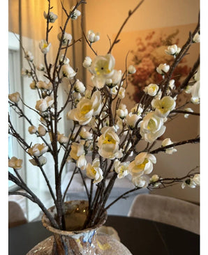 Un gros plan de la fleur de cerisier japonais artificielle de 94 cm avec de gracieuses fleurs de cerisier artificielles blanches au centre jaune sur des branches brunes dans un vase en verre, sur une table sombre avec une pièce élégante et doucement éclairée en arrière-plan.