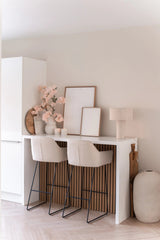 Un bar minimaliste avec deux tabourets beiges avec coussins, un comptoir blanc, des œuvres d'art encadrées, une lampe de table, une décoration aux tons neutres et une fleur de cerisier artificielle de 100 cm rose clair dans des vases contre un mur de couleur claire.