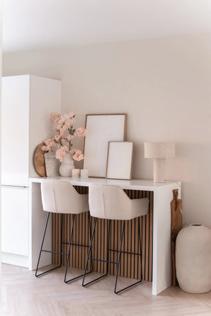 Un bar minimaliste avec deux tabourets beiges avec coussins, un comptoir blanc, des œuvres d'art encadrées, une lampe de table, une décoration aux tons neutres et une fleur de cerisier artificielle de 100 cm rose clair dans des vases contre un mur de couleur claire.