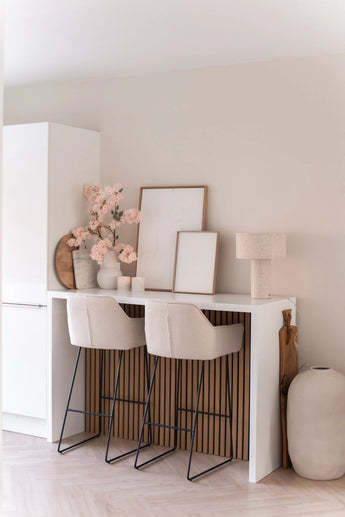 Un bar minimaliste avec deux tabourets beiges avec coussins, un comptoir blanc, des œuvres d'art encadrées, une lampe de table, une décoration aux tons neutres et une fleur de cerisier artificielle de 100 cm rose clair dans des vases contre un mur de couleur claire.
