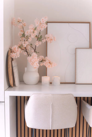 Un bureau minimaliste avec une chaise blanche, deux bougies, des plateaux décoratifs, des œuvres d'art abstraites encadrées et une fleur de cerisier artificielle de 100 cm rose clair dans un vase blanc sur une surface blanche.