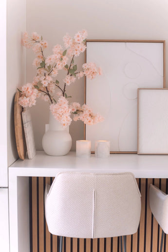 Un bureau minimaliste avec une chaise blanche, deux bougies, des plateaux décoratifs, des œuvres d'art abstraites encadrées et une fleur de cerisier artificielle de 100 cm rose clair dans un vase blanc sur une surface blanche.