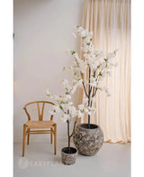 Une pièce minimaliste avec une chaise en bois, des rideaux beiges et deux fleurs de cerisier artificielles de 120 cm blanches avec des fleurs blanches dans un pot - l'une dans un pot gris avec structure et l'autre dans un pot noir.