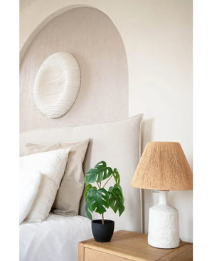 Un lit soigneusement fait avec du linge blanc se trouve à côté d'une table de chevet en bois avec l'Art Monstera 40 cm dans un pot décoratif noir et une lampe blanche avec un abat-jour tressé. Une arche douce et une œuvre d’art ronde et structurée sont accrochées au mur.