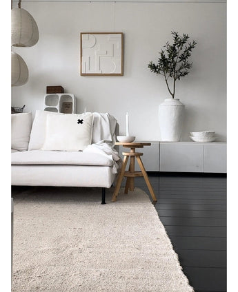 Een minimalistische woonkamer met een lichtgekleurde bank, neutraal vloerkleed, wit dressoir, Kunst Olijfboom 120 cm als decoratief element, abstracte muurkunst en houten krukjes-alles in zachte Mediterrane sfeer met neutrale tinten.