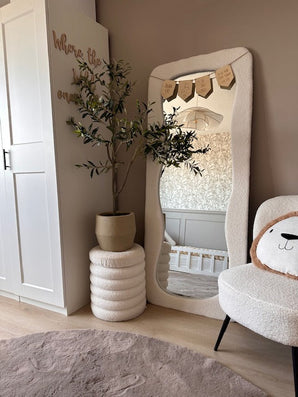 Een gezellige, moderne kinderkamer met neutrale tinten is voorzien van een golvende vloerspiegel, de Kunst Olijfboom 120 cm voor een mediterrane touch, een pluche stoel met een kussen met een berengezicht en een wieg die weerspiegeld wordt in de spiegel - de perfecte inrichting voor elke ruimte.