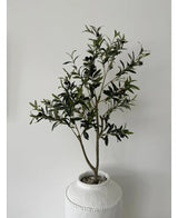 De Kunst Olijfboom 120 cm heeft groene bladeren en zwarte olijven in een witte pot met structuur en kiezels, waardoor het een prachtig decoratief element is tegen een effen lichtgrijze muur.