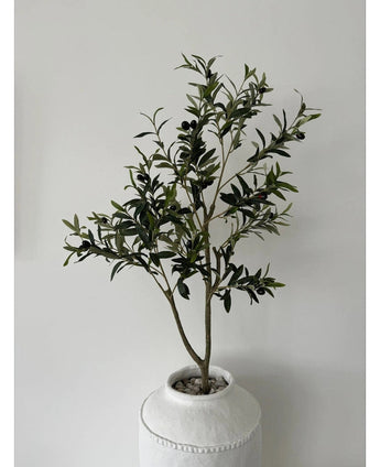 De Kunst Olijfboom 120 cm heeft groene bladeren en zwarte olijven in een witte pot met structuur en kiezels, waardoor het een prachtig decoratief element is tegen een effen lichtgrijze muur.