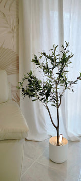 Een Kunst Olijfboom 120 cm met groene bladeren staat op een tegelvloer naast een crèmekleurige bank en geeft de kamer een Mediterrane sfeer, voor witte gordijnen en een muur met subtiel patroon.