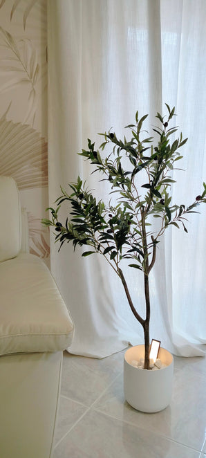 Een Kunst Olijfboom 120 cm met groene bladeren staat op een tegelvloer naast een crèmekleurige bank en geeft de kamer een Mediterrane sfeer, voor witte gordijnen en een muur met subtiel patroon.