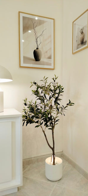 Een Kunst Olijfboom 120 cm met groene bladeren staat in de hoek van een lichte kamer en brengt een mediterraan tintje. Erboven hangen twee ingelijste kunstprints van vazen met takken, terwijl een witte kast en lamp gedeeltelijk zichtbaar zijn aan de linkerkant.