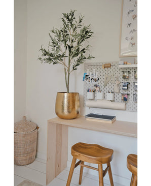 Een moderne werkplek met een houten bureau, twee krukjes, een grote gouden plantenbak met de Kunst Olijfboom 120 cm voor een mediterrane flair, een wasbord met kantoorbenodigdheden en een rieten mand op de vloer als decoratief detail.