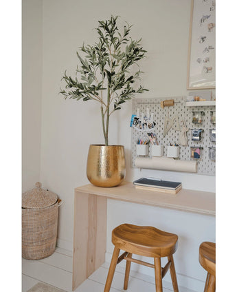 Een moderne werkplek met een houten bureau, twee krukjes, een grote gouden plantenbak met de Kunst Olijfboom 120 cm voor een mediterrane flair, een wasbord met kantoorbenodigdheden en een rieten mand op de vloer als decoratief detail.