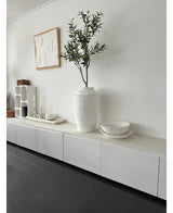 Een minimalistische kamer met een wit dressoir, de Kunst Olijfboom 120 cm als decoratief element, decoratieve witte schalen, een kaars en abstracte wandkunst. Donkerhouten vloeren en moderne accenten creëren een serene Mediterrane sfeer.
