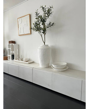 Een minimalistische kamer met een wit dressoir, de Kunst Olijfboom 120 cm als decoratief element, decoratieve witte schalen, een kaars en abstracte wandkunst. Donkerhouten vloeren en moderne accenten creëren een serene Mediterrane sfeer.