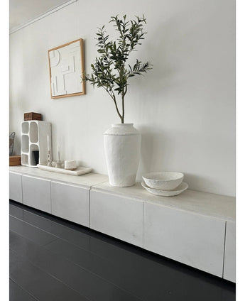 Een minimalistische kamer met een wit dressoir, de Kunst Olijfboom 120 cm als decoratief element, decoratieve witte schalen, een kaars en abstracte wandkunst. Donkerhouten vloeren en moderne accenten creëren een serene Mediterrane sfeer.
