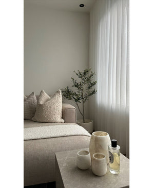 Een minimalistische woonkamer heeft een beige bank, structuurkussens, een Kunst Olijfboom 120 cm als decor, transparante witte gordijnen en een marmeren tafel met drie crèmekleurige vazen en parfum. Natuurlijk licht verlicht de rustige ruimte.