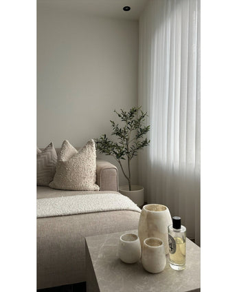 Een minimalistische woonkamer heeft een beige bank, structuurkussens, een Kunst Olijfboom 120 cm als decor, transparante witte gordijnen en een marmeren tafel met drie crèmekleurige vazen en parfum. Natuurlijk licht verlicht de rustige ruimte.