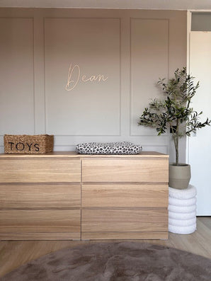 Een minimalistische kinderkamer bevat een houten commode met een aankleedkussen met luipaardprint, een geweven mand met het label "Toys" als decoratie, de Kunst Olijfboom 120 cm voor groen en "Dean" in cursief op de beige muur erboven.