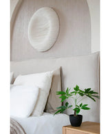 Un lit soigneusement fait avec une literie blanche se trouve à côté d'une table de chevet en bois avec la plante artificielle Pachira de 30 cm. Au-dessus du lit, une peinture murale circulaire structurée blanche décore le mur de couleur claire.