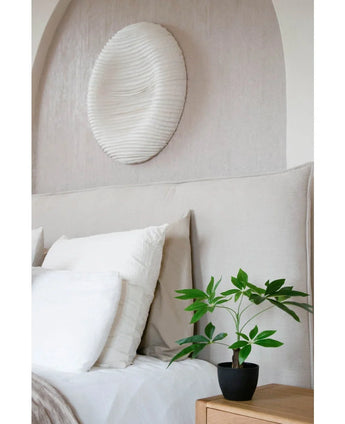 Un lit soigneusement fait avec une literie blanche se trouve à côté d'une table de chevet en bois avec la plante artificielle Pachira de 30 cm. Au-dessus du lit, une peinture murale circulaire structurée blanche décore le mur de couleur claire.