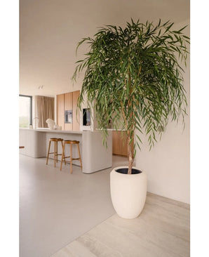Un saule artificiel de 210 cm aux feuilles longues et minces ajoute de la verdure à une cuisine moderne avec des armoires en bois clair, un îlot blanc et des tabourets de bar en bois. La lumière du soleil provenant de la fenêtre souligne sa présence dans cet espace minimaliste.