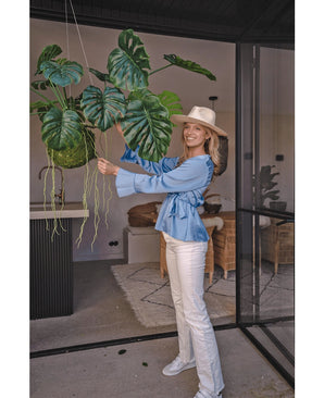 Une femme souriante avec un chapeau, un chemisier bleu et un pantalon blanc se tient à l'intérieur et tient une plante artificielle suspendue Monstera Deluxe 60 cm avec de grandes feuilles vertes, entourée d'un décor moderne pour une ambiance tropicale avec des plantes décoratives.