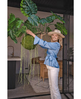 Une femme souriante vêtue d'un chemisier bleu, d'un pantalon blanc et d'un chapeau montre la plante artificielle suspendue Monstera Deluxe 80 cm avec des vignes vertes sinueuses, qui apporte une atmosphère tropicale à l'espace intérieur lumineux avec des murs en verre et des meubles en rotin.