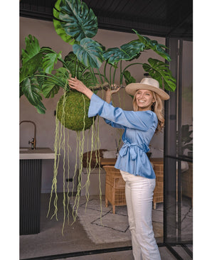 Une femme souriante vêtue d'un chemisier bleu, d'un pantalon blanc et d'un chapeau montre la plante artificielle suspendue Monstera Deluxe 80 cm avec des vignes vertes sinueuses, qui apporte une atmosphère tropicale à l'espace intérieur lumineux avec des murs en verre et des meubles en rotin.