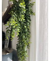 Un gros plan de la plante artificielle suspendue Pancake 61 cm, une plante à feuilles vertes rampantes suspendue à une planche de bois près d'un rideau blanc translucide, avec une partie d'un vase gris foncé en dessous.
