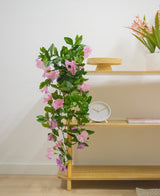 Une étagère en bois avec plante artificielle suspendue Pétunia 70 cm violet, une horloge blanche, un support en bois, un livre et un vase avec des fleurs roses contre un mur blanc.