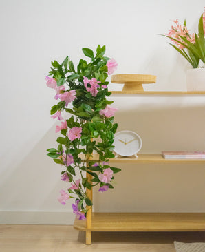 Une étagère en bois avec plante artificielle suspendue Pétunia 70 cm violet, une horloge blanche, un support en bois, un livre et un vase avec des fleurs roses contre un mur blanc.
