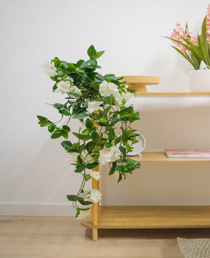 La plante artificielle suspendue Pétunia 70 cm blanche avec des fleurs blanches tombe d'une étagère en bois dans une pièce lumineuse et moderne avec un parquet en bois clair. Sur l'étagère du haut se trouve un vase avec de longues fleurs roses.