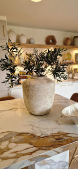Un vase beige rustique avec une feuille d'olivier artificielle de 90 cm ignifuge se dresse sur un comptoir en marbre dans une cuisine moderne et chaleureusement éclairée, avec des étagères et une décoration méditerranéenne en arrière-plan.