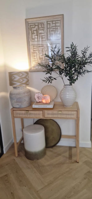 Une console en bois avec deux tiroirs contient une lampe, un grand vase blanc avec feuille d'olivier artificielle de 90 cm ignifuge, un plus petit vase, des livres et des pierres décoratives. Des accents méditerranéens et des œuvres d'art abstraites encadrées complètent le look ci-dessus. Au fond se trouvent un tabouret et un plateau rond.